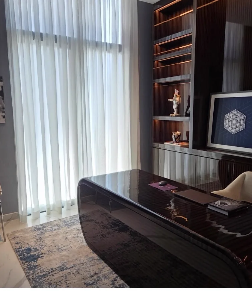 Office with a dark sculptural desk, bespoke cabinetry with integrated lighting and a tall curtained window — Contemporary International Villa